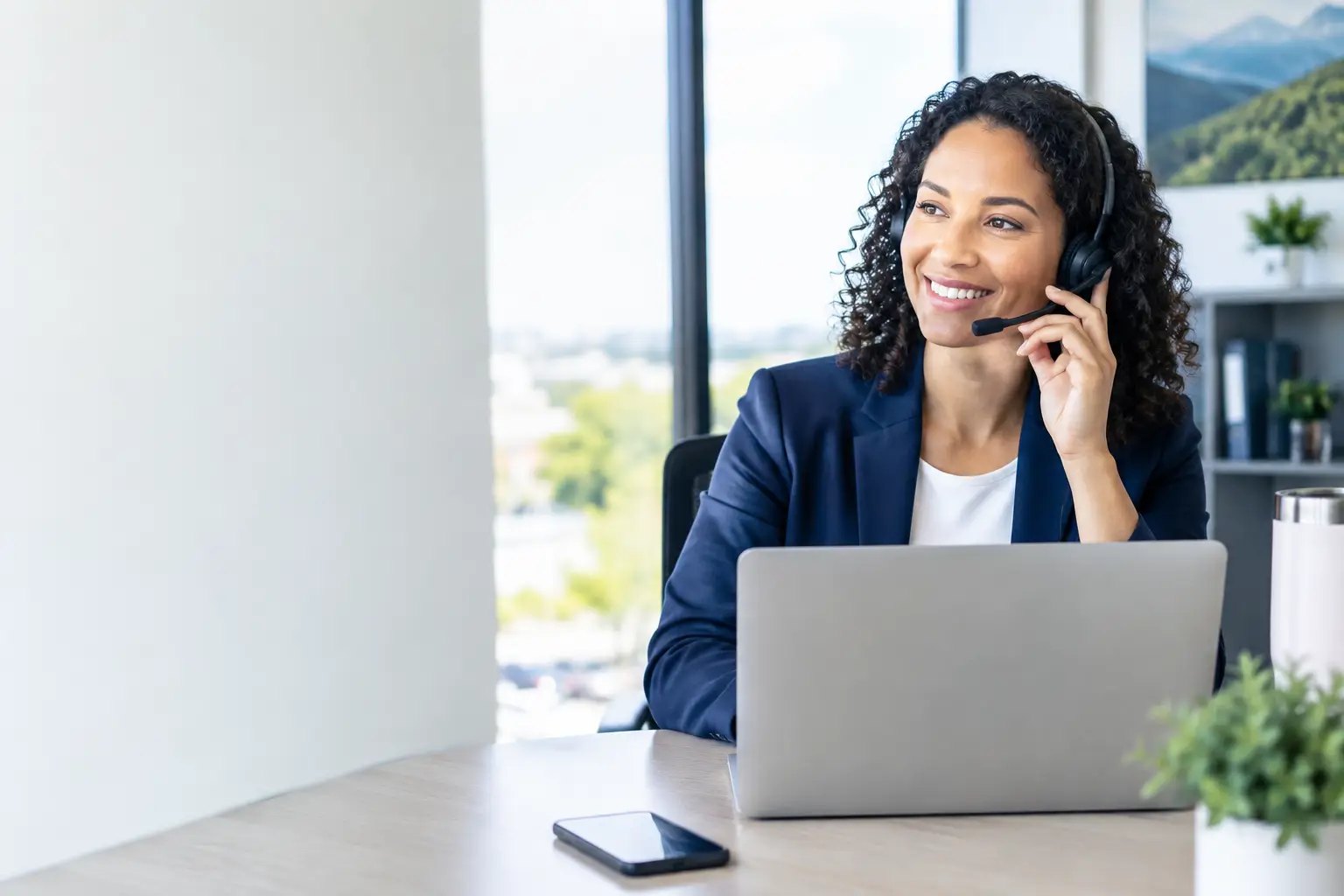 Smiling office professional wearing a headset at a laptop in a bright office, answering a call on her business phone number