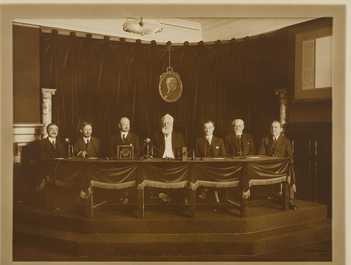 Alexander Graham Bell, white bearded and seated at the center of a long draped table with six AT&T officials, at the January 25, 1915 opening of the first transcontinental telephone line in New York