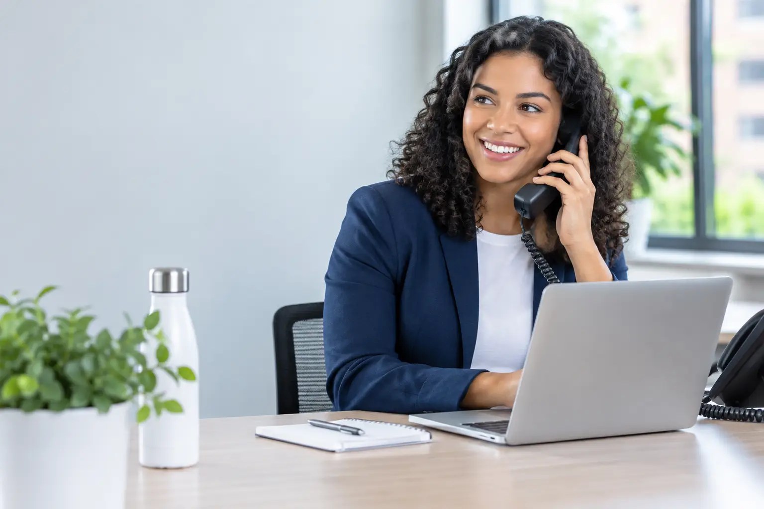 Smiling office manager in a navy blazer answering a desk phone while working at her laptop