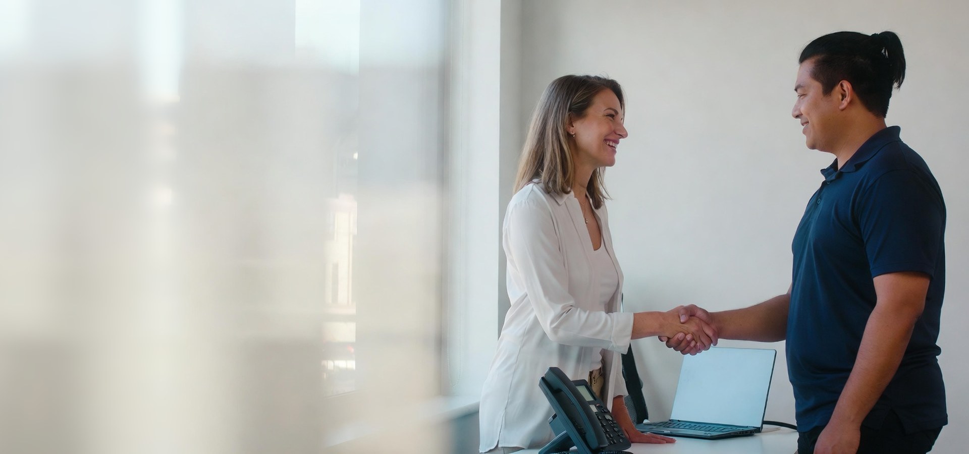 Small business owner shaking hands with a local phone technician in a bright office, with a desk phone and laptop on the desk beside them