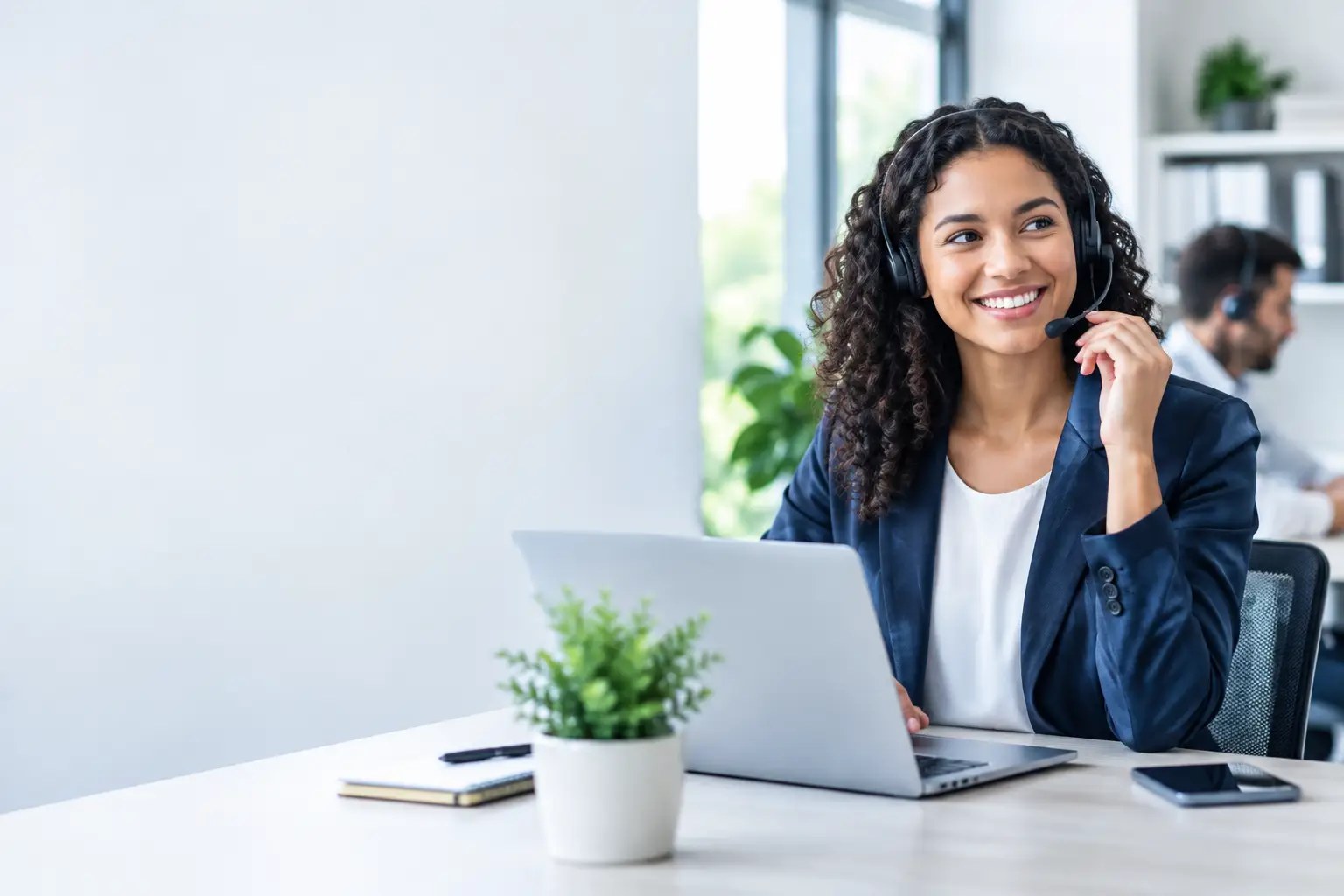 Smiling office professional wearing a headset at her laptop, looking toward a bright window, with a coworker on a call in the background