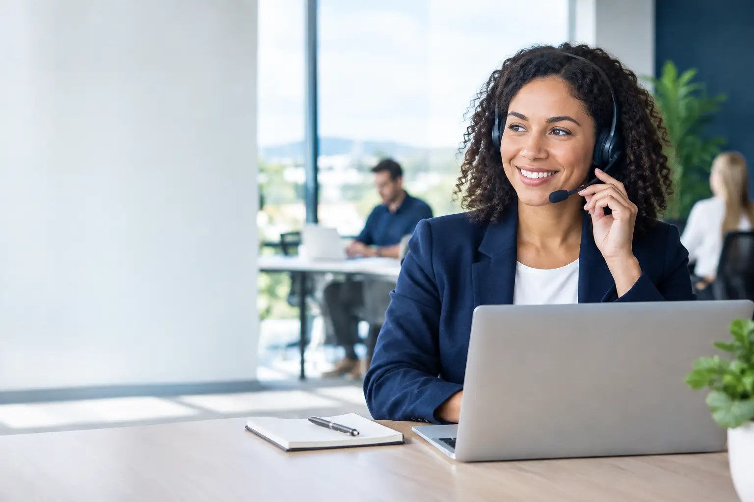 Receptionist wearing a headset and smiling at her laptop in a bright office, with two coworkers at desks behind her