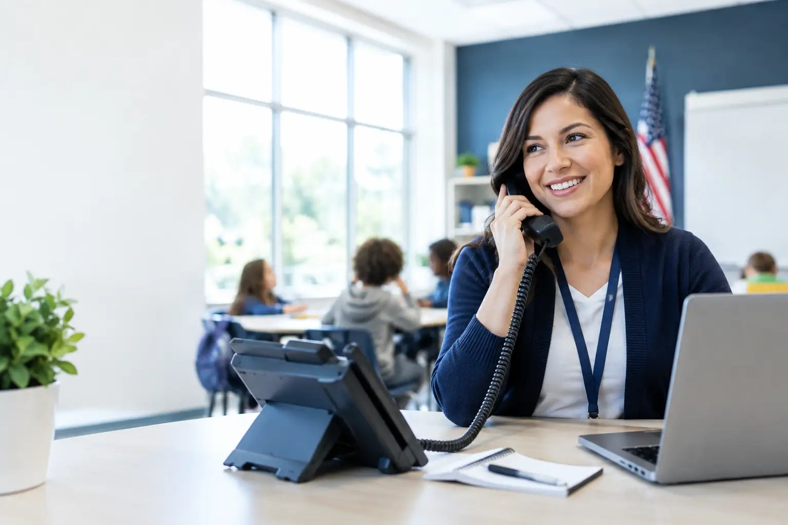 School staff member smiling on a desk phone in a classroom, with students at tables and an American flag behind her