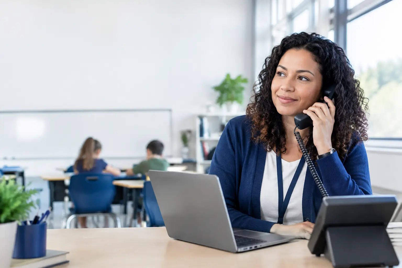 Teacher on a classroom desk phone with students working at desks behind her, illustrating E911 for schools