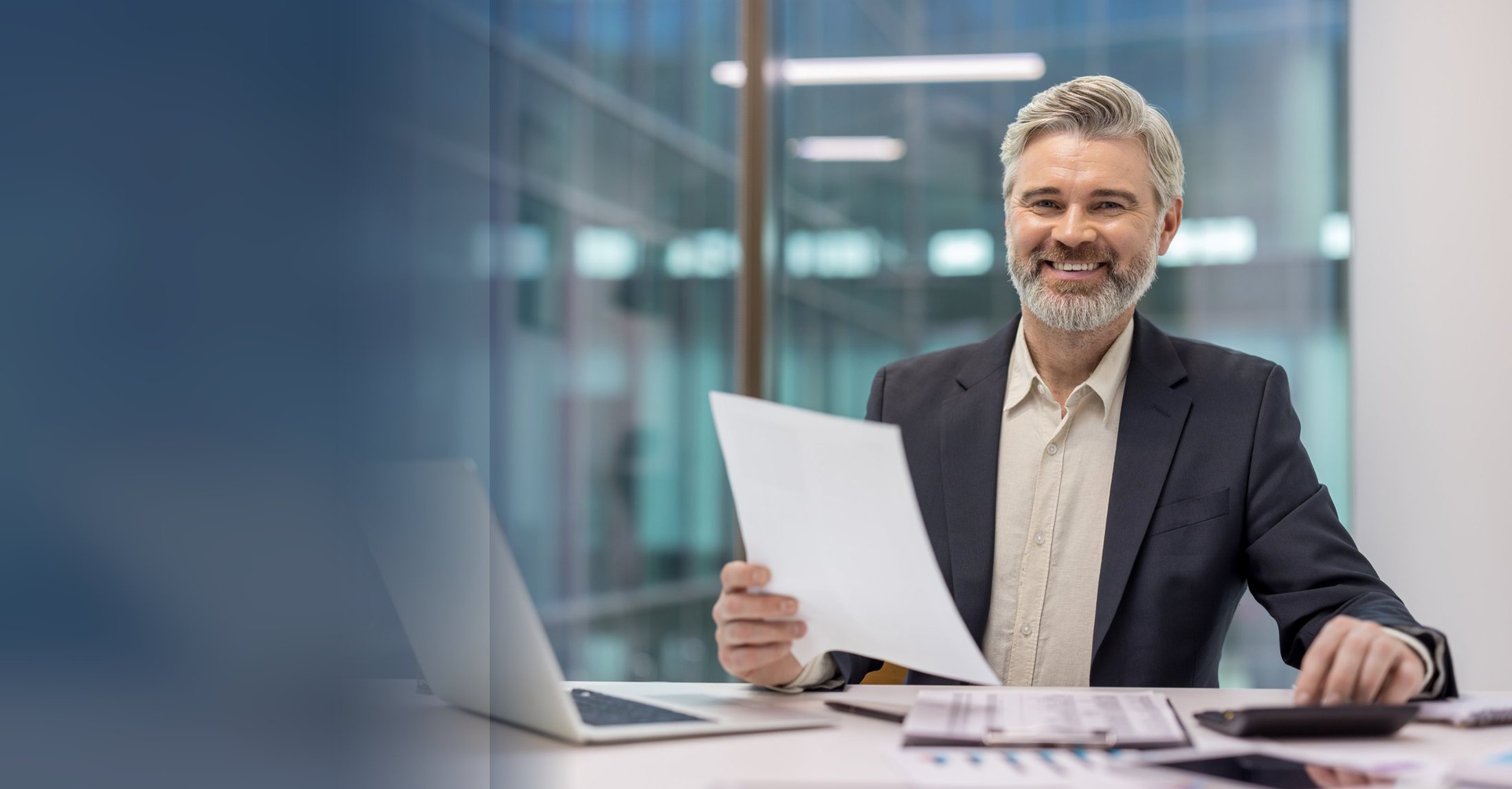 A financial advisor at his desk holding a document, with a laptop and a calculator beside him