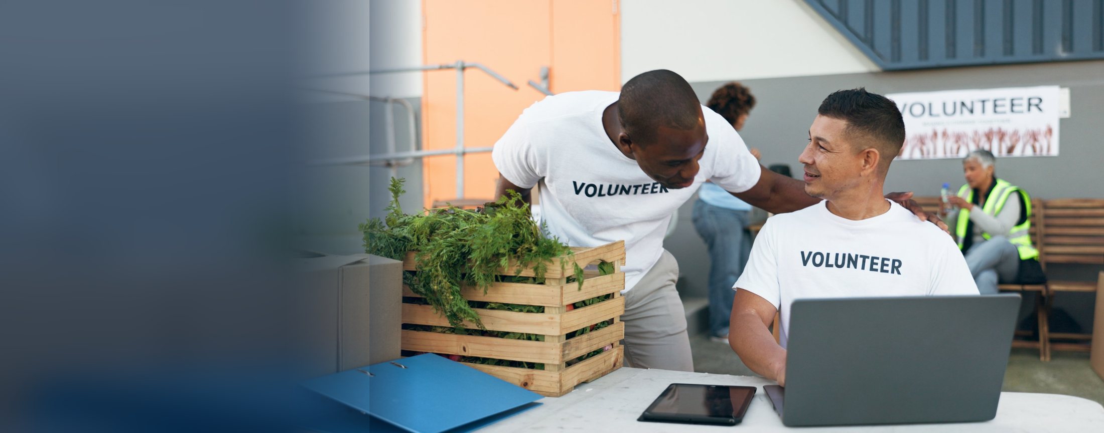 Two volunteers talking at a food drive table beside a crate of fresh produce and a laptop